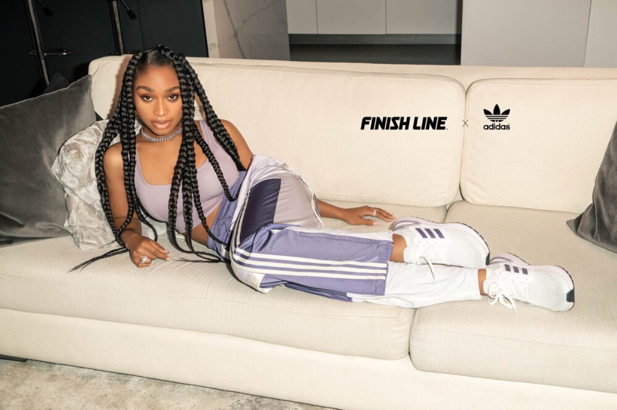 Woman in athletic pants and crop top poses on a white couch with white sneakers. Brands: Adidas and Finish Line are visible.