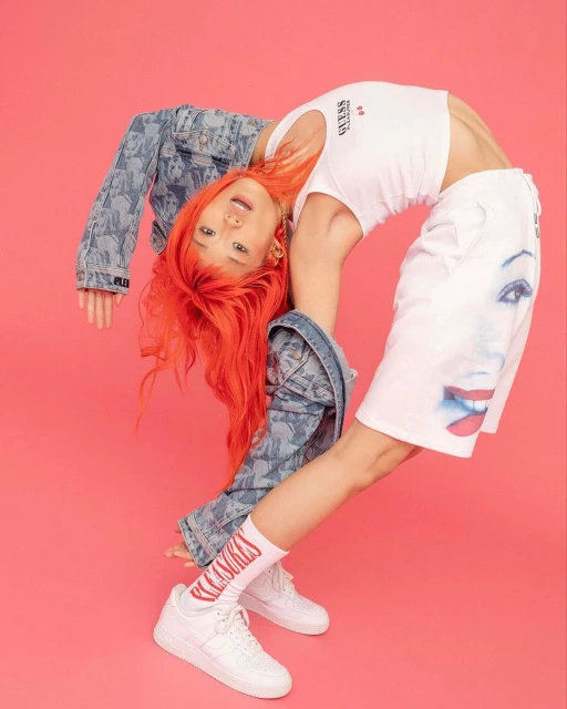 Young woman with red hair bends over backwards in studio, wearing a white top, shorts with face graphic, denim jacket, socks and sneakers.