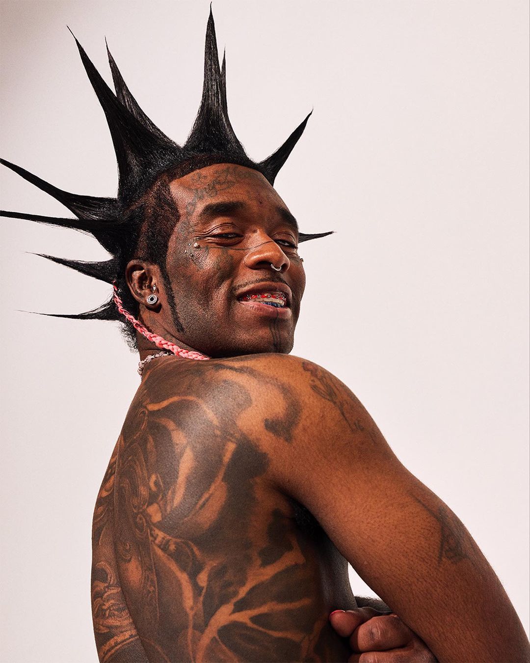 Close-up studio shot of an adult Black male model with a mohawk hairstyle, tattoos on his back, and jewelry, against a plain white background.