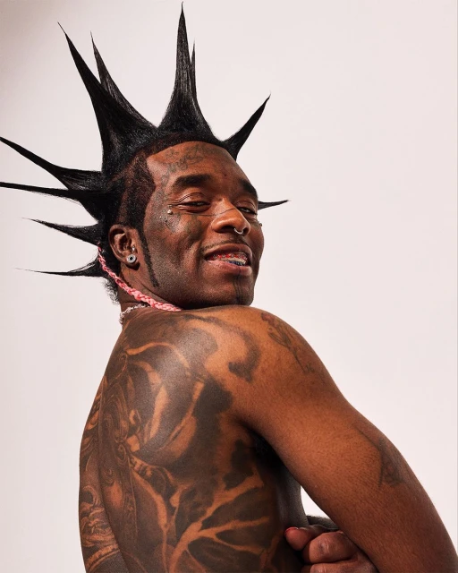 Close-up studio shot of an adult Black male model with a mohawk hairstyle, tattoos on his back, and jewelry, against a plain white background.