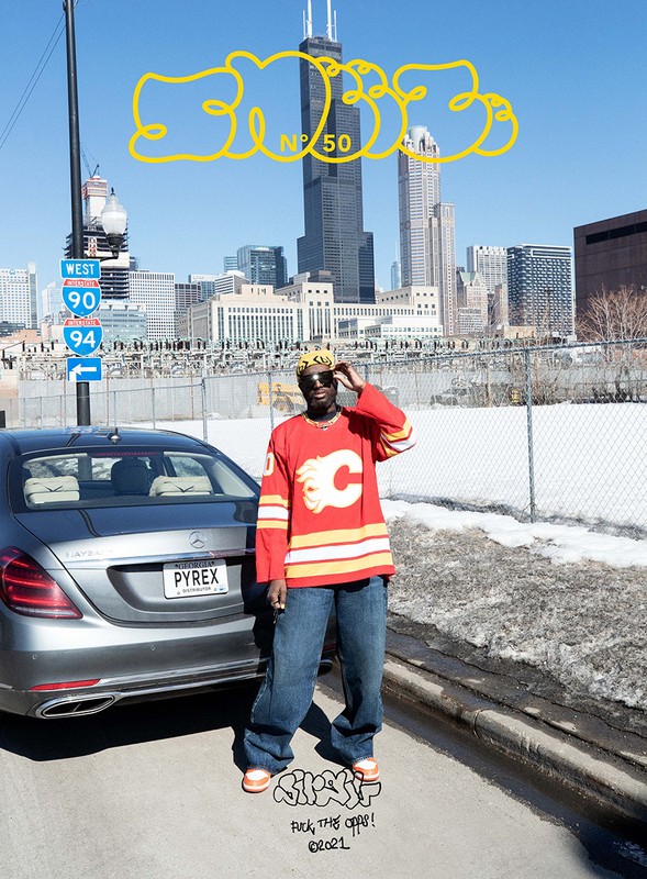 Man in sports jersey poses near car in urban setting with Chicago skyline and overcast sky in the background on a sunny day.