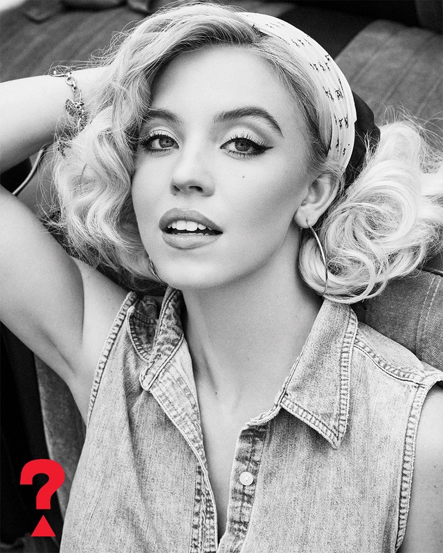 Black-and-white portrait of a young woman with curly hair and a headscarf, wearing a denim top, posing against a draped background.