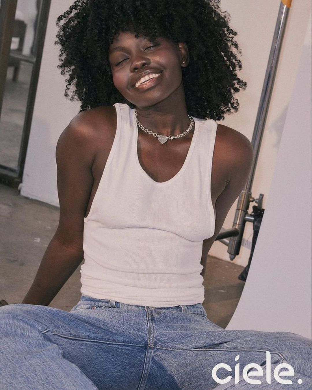 Portrait of a smiling Black woman with curly hair wearing a white tank top and silver necklace, photographed by Kenneth Cappello.