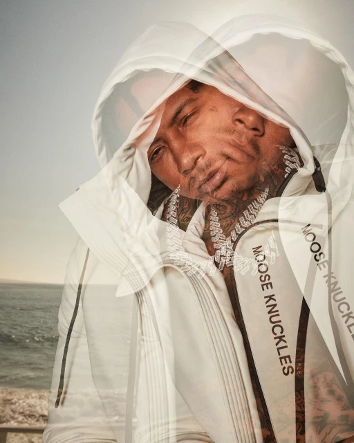 Portrait of an adult male wearing a white hooded sweatshirt with 'MOOSE KNUCKLES' text, set against an ocean backdrop, showcasing fashion apparel.