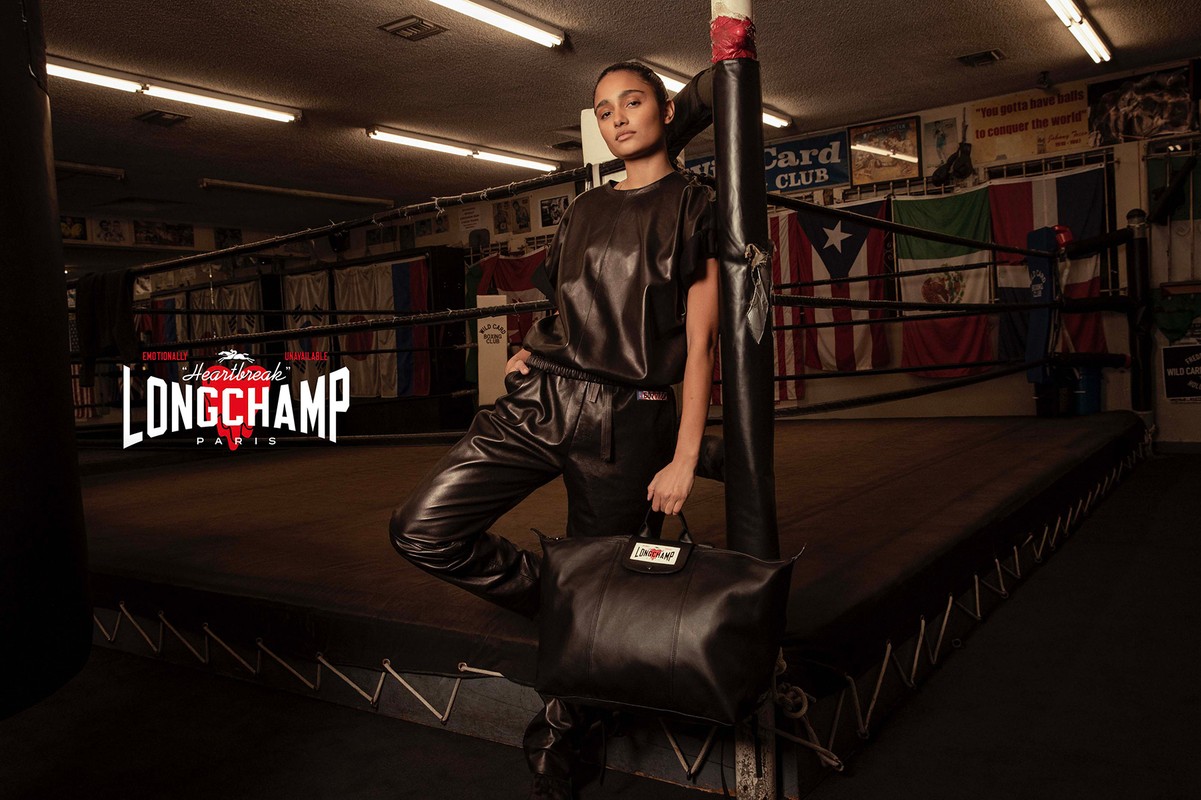 Adult female fashion model poses with a black leather handbag inside a boxing ring wearing black leather top and pants for Longchamp.