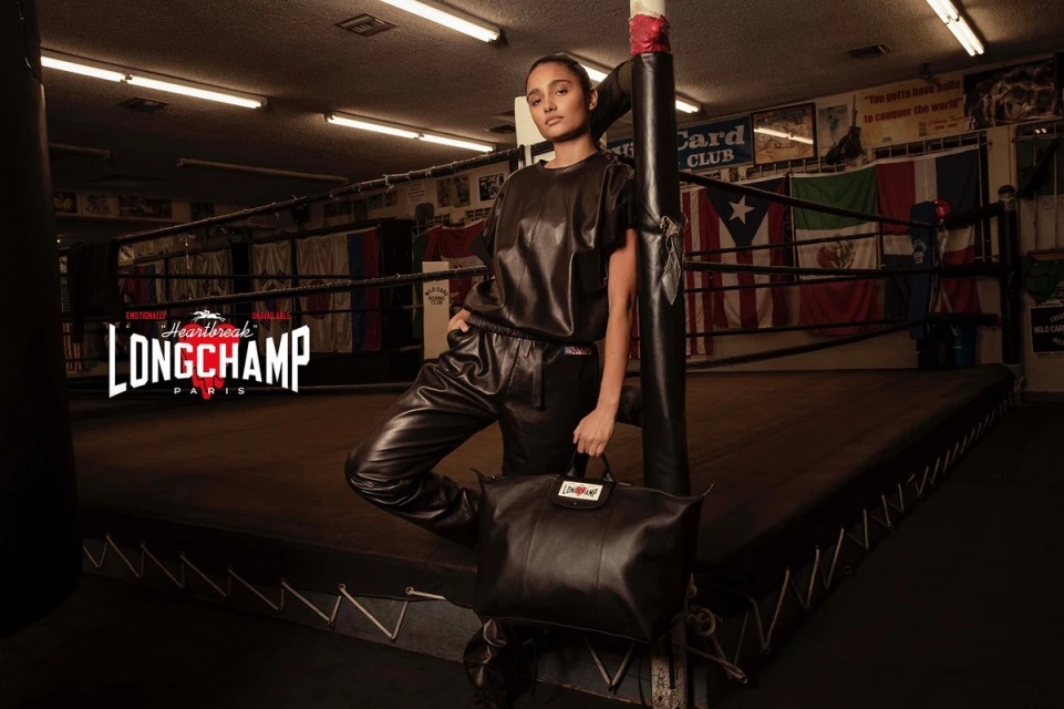 Adult female fashion model poses with a black leather handbag inside a boxing ring wearing black leather top and pants for Longchamp.
