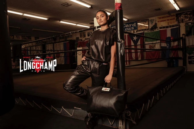 Adult female fashion model poses with a black leather handbag inside a boxing ring wearing black leather top and pants for Longchamp.