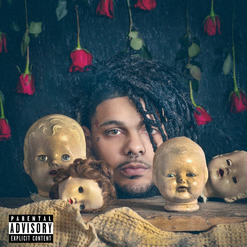 Music album cover featuring a male musician with dreadlocks surrounded by antique dolls and red roses in a dark and moody setting.