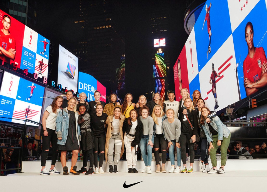 Group portrait of US women's soccer team in Times Square at night surrounded by digital billboards displaying soccer players.