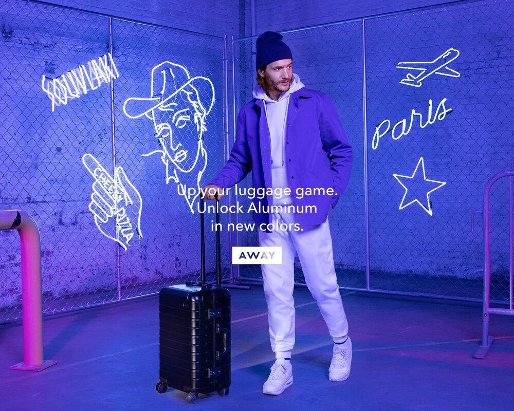 A man in a purple jacket stands next to black rolling luggage in a neon-lit indoor setting promoting modern travel accessories.
