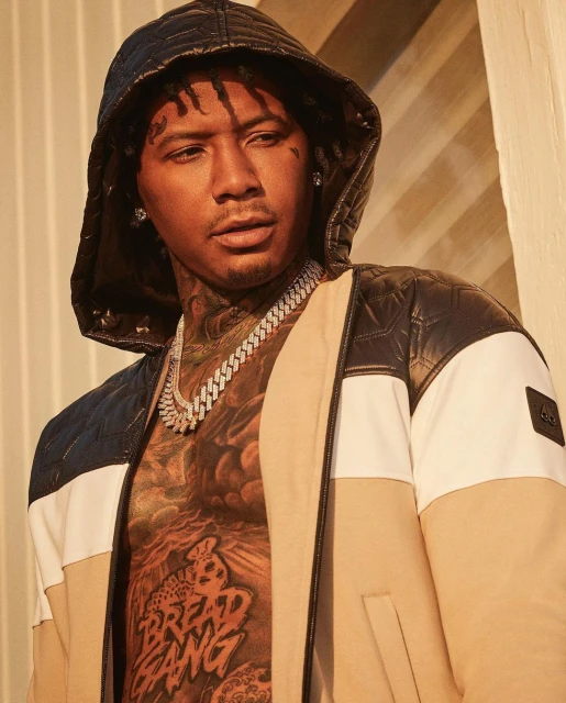 Close-up of a tattooed adult male wearing a hooded jacket and diamond chain necklace, posing indoors in a photo studio setting.