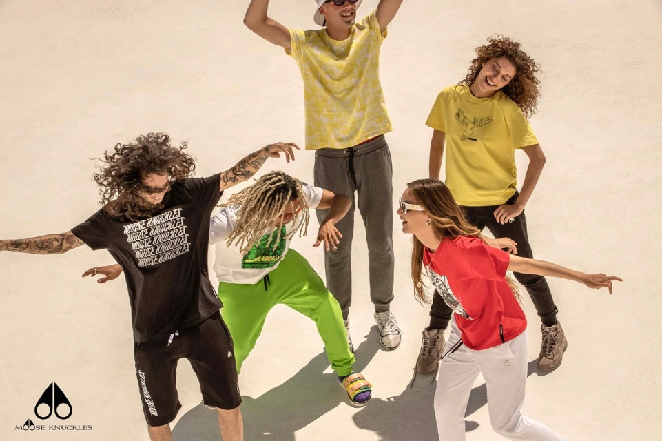 Group of five diverse adults in fashionable streetwear, posing and interacting dynamically against a light backdrop, showcasing style and brand.