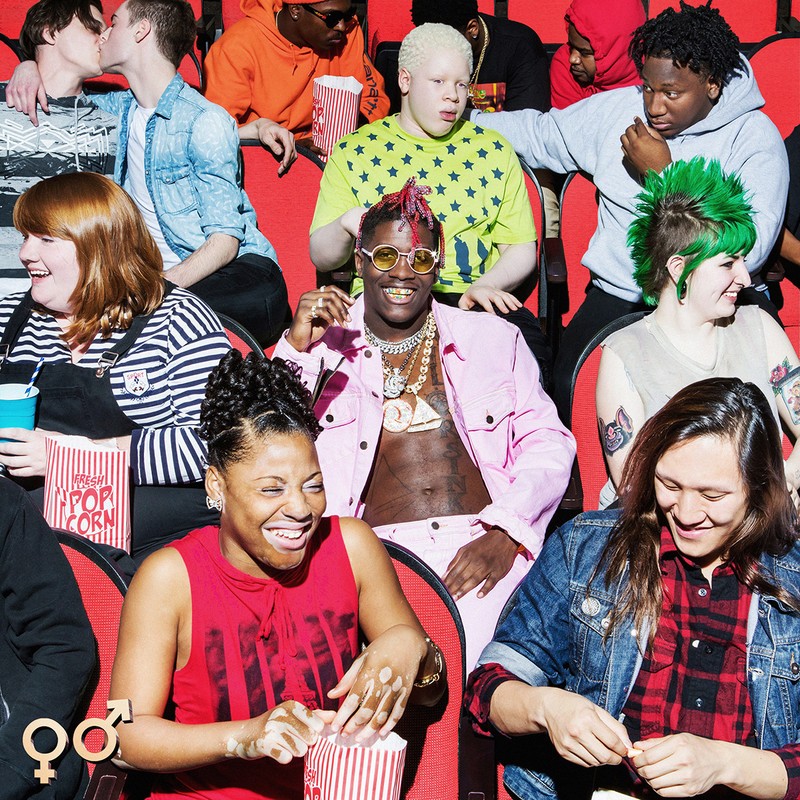 Group of diverse adults in a movie theatre, eating popcorn, and laughing together. A man with pink hair sits prominently in the middle.