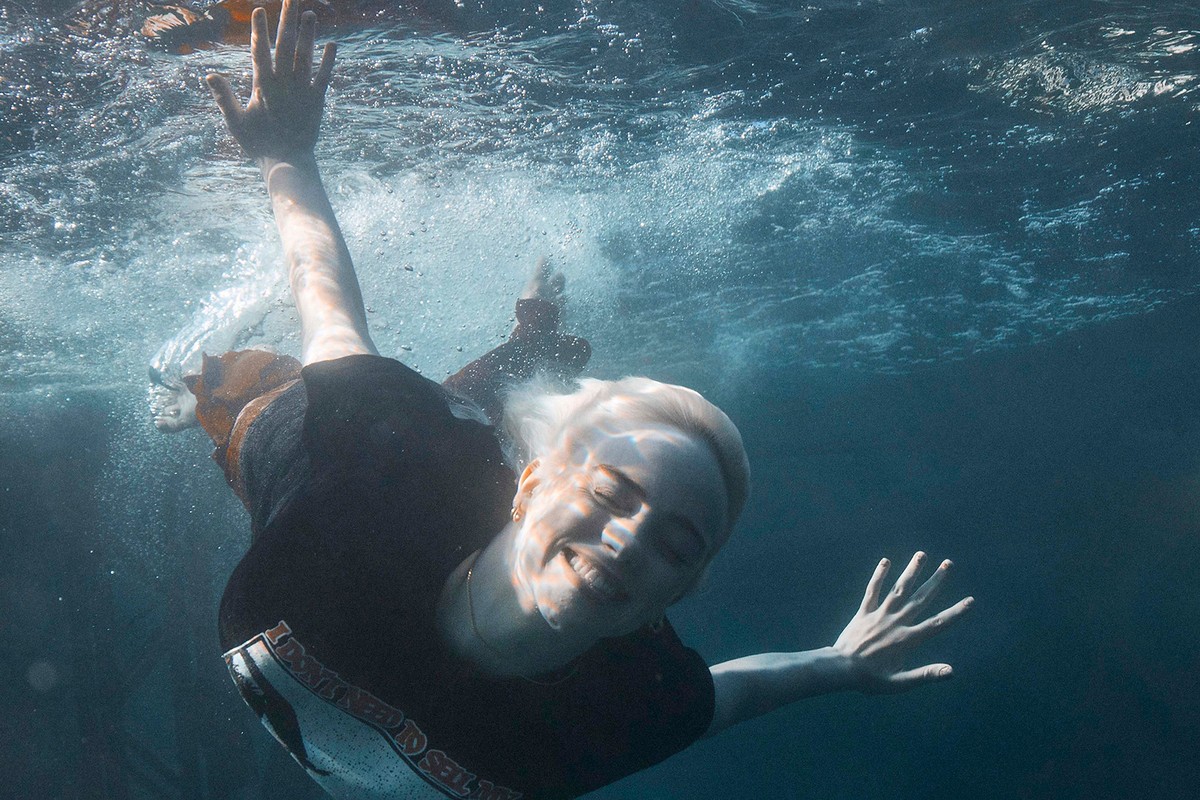 Woman with blond hair swims underwater in a black top, smiling and looking directly at the camera with arms outstretched in clear blue water.