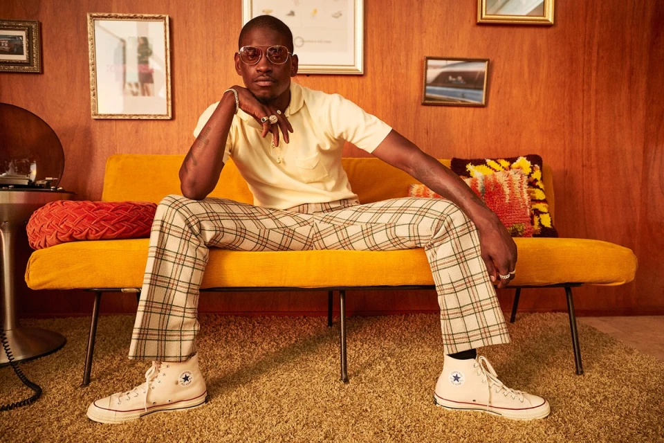 An African American man in sunglasses and plaid pants sits on a yellow couch indoors with pictures on the paneled walls behind him.