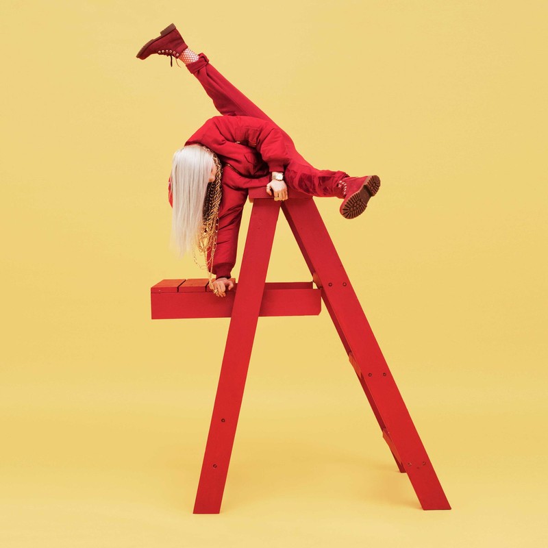 Billie Eilish strikes a dynamic pose on a red lifeguard chair against a yellow backdrop, showcasing her edgy style and acrobatic ability.