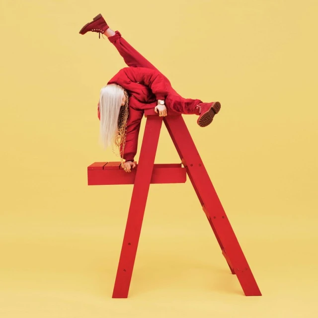 Billie Eilish strikes a dynamic pose on a red lifeguard chair against a yellow backdrop, showcasing her edgy style and acrobatic ability.