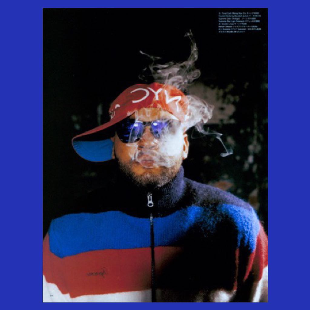 Man with a beard exhales cigarette smoke wearing mirrored sunglasses, a cap, and a color-blocked jacket in a close-up studio portrait.