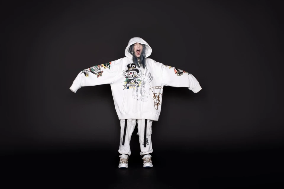 A person stands facing forward with arms outstretched, wearing a white hooded jacket and pants against a dark gray backdrop.