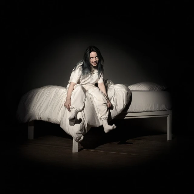 Billie Eilish sits on a bed in a white outfit, smiling with dark, teal-tinted hair in a dark studio with wood floors and shadows.