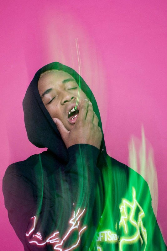 Portrait of a young adult with gold teeth wearing a hooded sweatshirt in front of a pink background with green lighting.