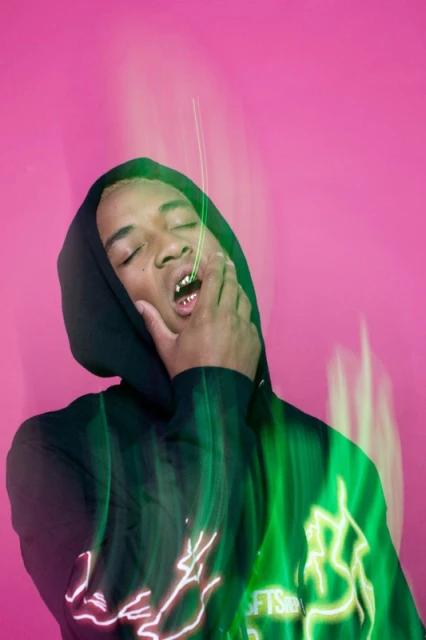Portrait of a young adult with gold teeth wearing a hooded sweatshirt in front of a pink background with green lighting.