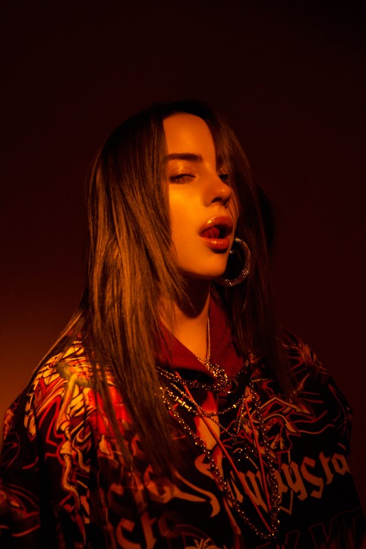Portrait of Billie Eilish with dark hair, hoop earrings, layered necklaces, and a patterned top under warm amber lighting.