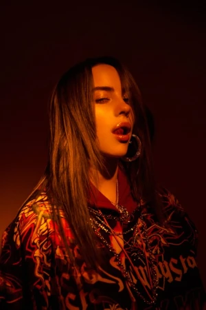 Portrait of Billie Eilish with dark hair, hoop earrings, layered necklaces, and a patterned top under warm amber lighting.