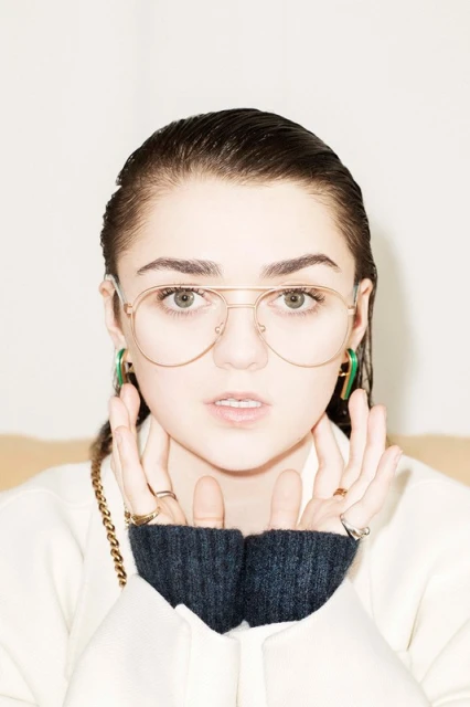 Close-up studio portrait of a woman with slicked-back hair wearing gold aviator glasses, rings, and statement earrings with hands raised.