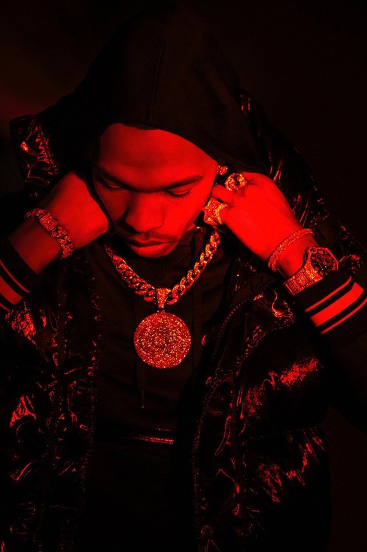 Man wearing gold jewelry and a hooded jacket is illuminated by red lighting in a studio portrait, showcasing fashion and style.