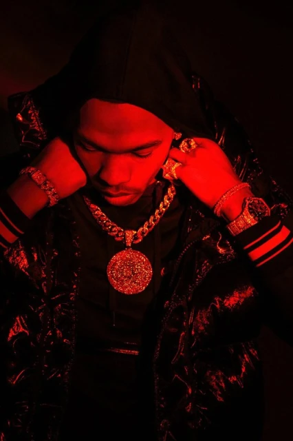 Man wearing gold jewelry and a hooded jacket is illuminated by red lighting in a studio portrait, showcasing fashion and style.