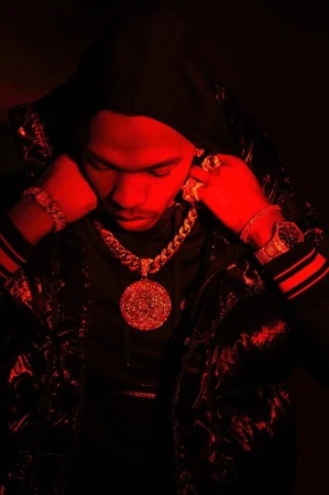 Man wearing gold jewelry and a hooded jacket is illuminated by red lighting in a studio portrait, showcasing fashion and style.