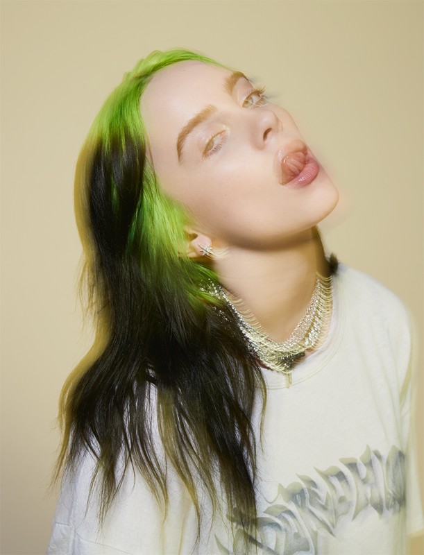 Billie Eilish with bright green hair roots and dark lengths, sticking out tongue, wearing white t-shirt and layered necklace against warm background