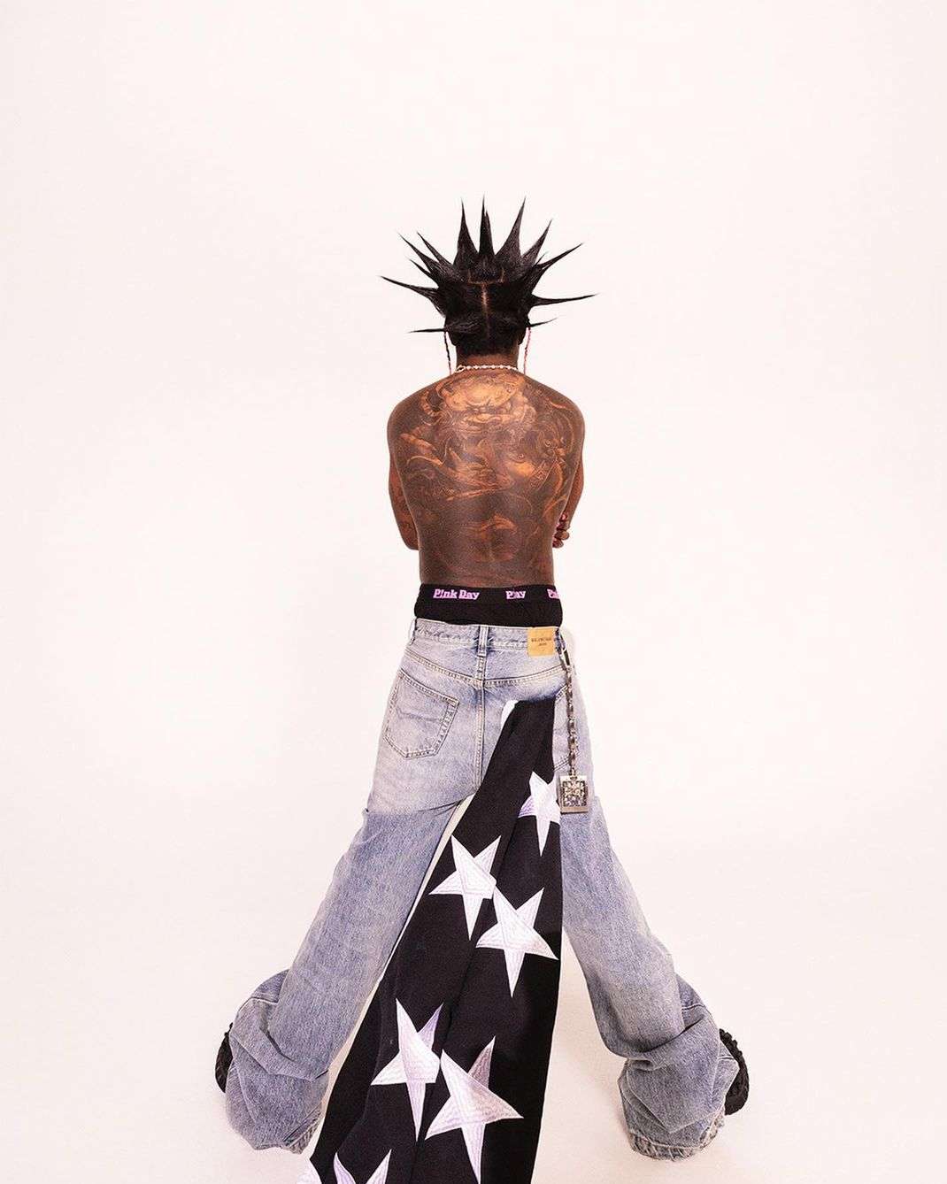 Person with mohawk, tattoos, and draped flag poses in light-wash denim jeans against a bright white backdrop in an eye-level shot.