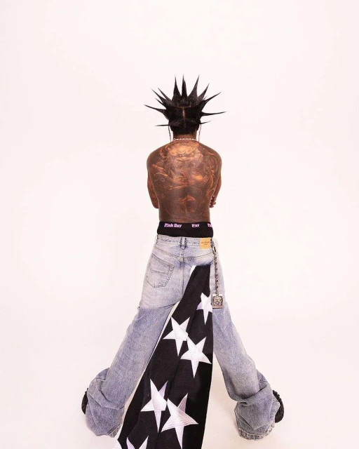 Person with mohawk, tattoos, and draped flag poses in light-wash denim jeans against a bright white backdrop in an eye-level shot.
