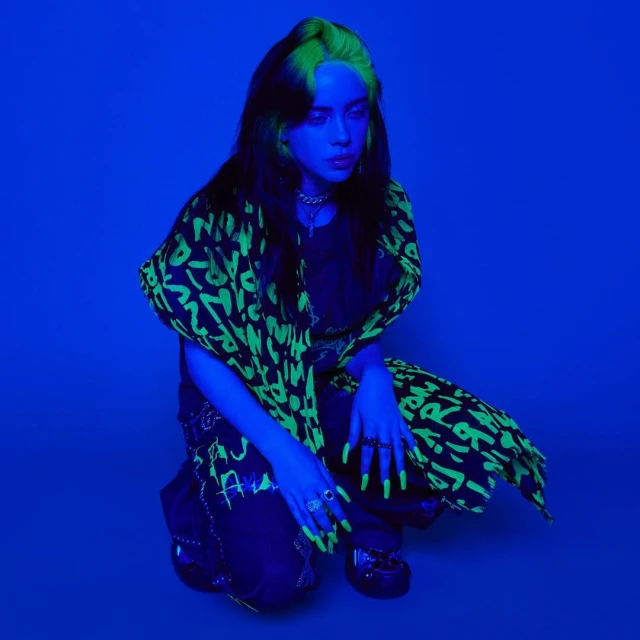 Billie Eilish in a blue-lit photo studio, crouching and wearing a black and neon green graphic patterned jacket and pants with chunky rings.