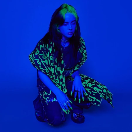 Billie Eilish in a blue-lit photo studio, crouching and wearing a black and neon green graphic patterned jacket and pants with chunky rings.