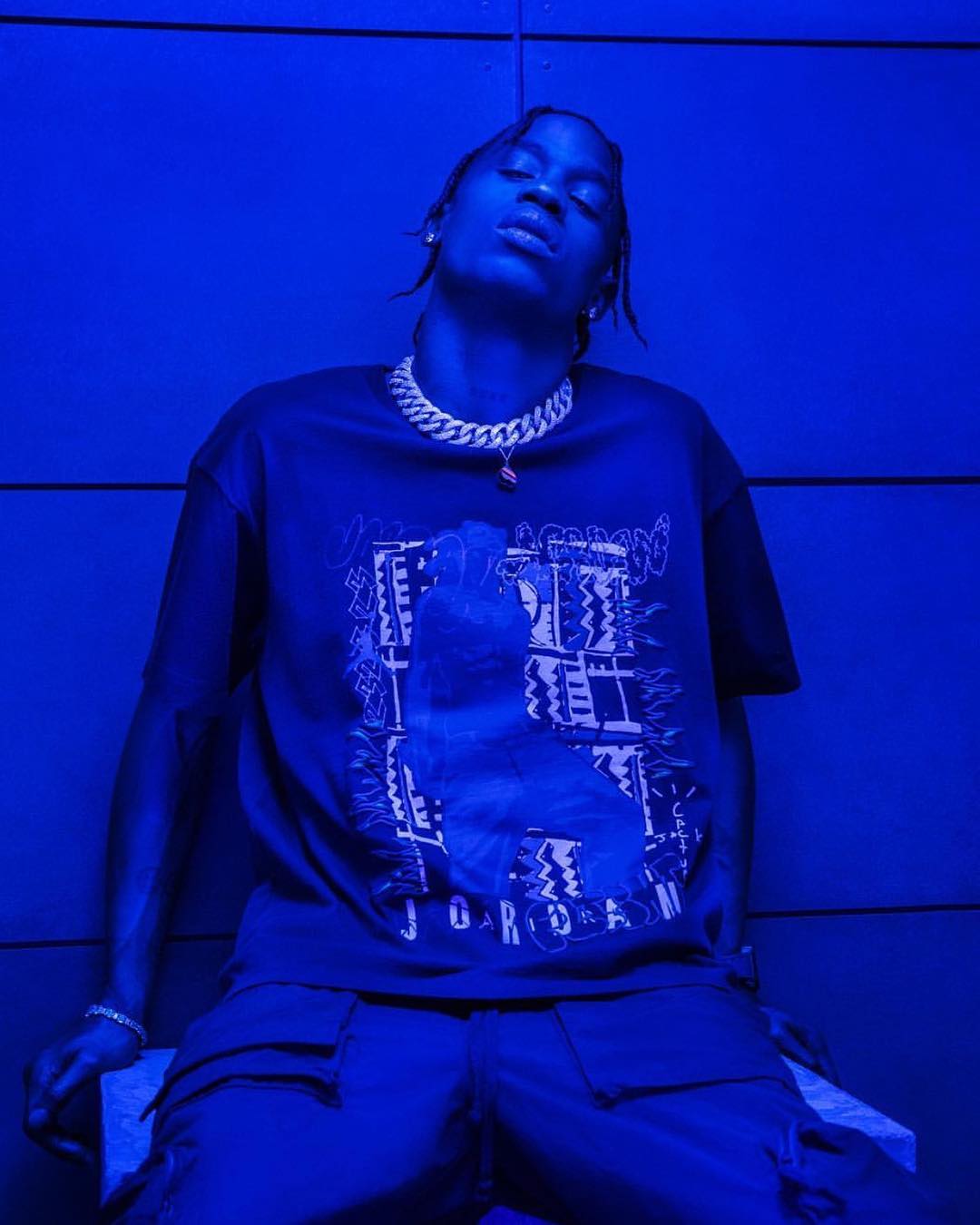 Young adult wearing a graphic t-shirt and jewelry, posing in an indoor blue-lit setting against a textured blue wall.