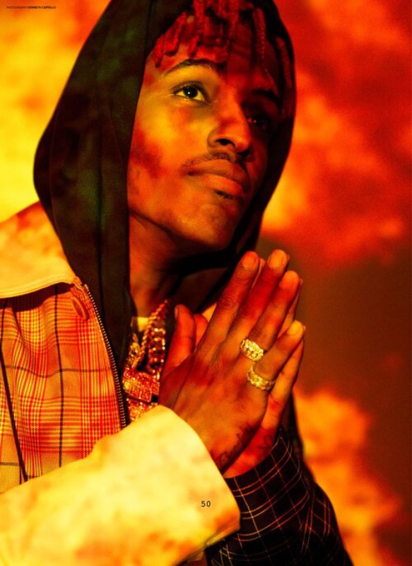 Portrait of a young man with dreadlocks, wearing a hoodie and plaid jacket, gold jewelry, hands together in front of a red and orange background.