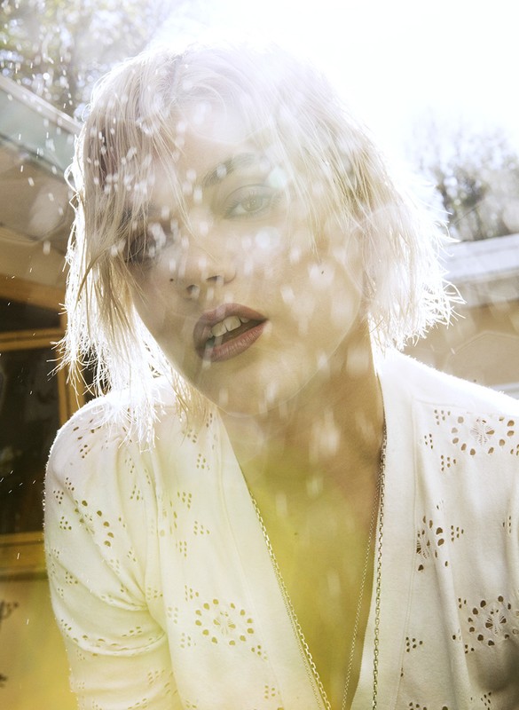 Close-up shot of a fair-skinned woman with blonde hair wearing a white eyelet top and silver necklace in a sun-drenched outdoor setting.