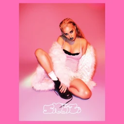 Blonde woman in pink top and fluffy white coat crouching against a pink studio background