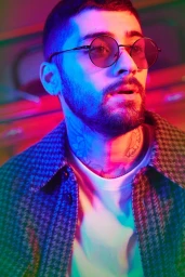 Close-up portrait of Zayn Malik wearing round tinted sunglasses, a white t-shirt, and a patterned jacket under vibrant neon lighting.
