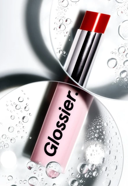Close-up studio shot of pink Glossier lip balm with red cap, reflective surfaces, and water droplets, highlighting the cosmetic product.