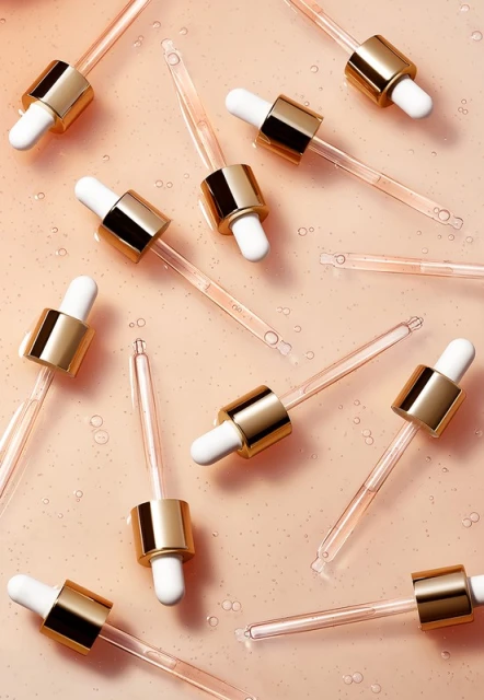 Multiple clear glass droppers with gold accents are arranged on a smooth peach surface with droplets of liquid for beauty product imagery.
