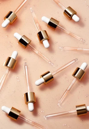 Multiple clear glass droppers with gold accents are arranged on a smooth peach surface with droplets of liquid for beauty product imagery.