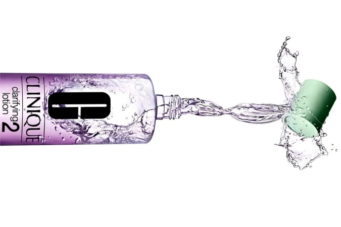 Clinique clarifying lotion bottle lying horizontally with cap off and liquid splashing. Product shot on a white background.