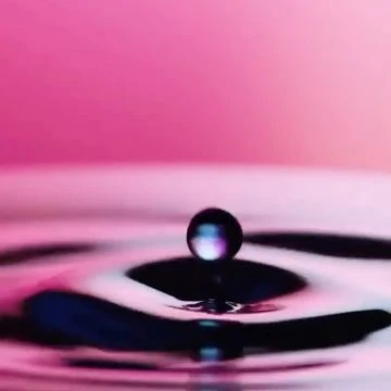High-speed photograph of a water drop impacting a smooth water surface, creating ripples against a pink-to-purple gradient background.