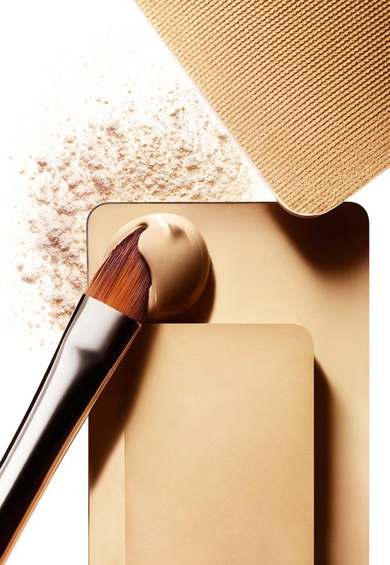 Tan foundation on a makeup brush atop a stack of cosmetic compacts and loose powder in a well-lit, neutral-toned beauty still life.