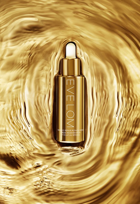 Gold bottle of Eve Lom skincare product is centered on a background of moving gold liquid, creating a luxurious beauty product shot.