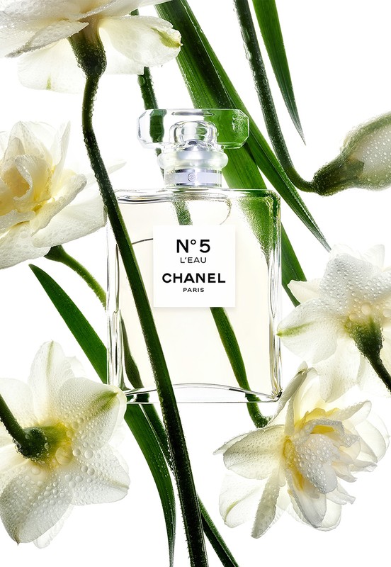 Close-up of a Chanel No. 5 L'Eau perfume bottle surrounded by white daffodils with water droplets and green stems against a white background.